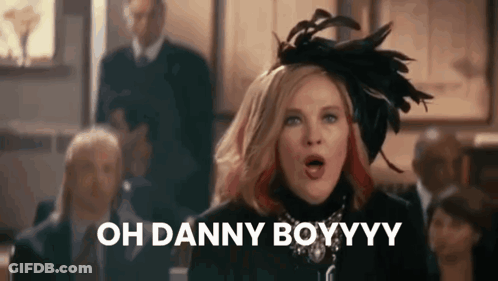 A Woman Wearing A Feathered Hat Is Standing In Front Of A Group Of People And Saying Oh Danny Boyyyy . GIF