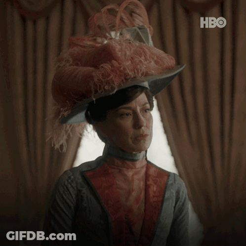 A Woman Wearing A Feathered Hat Says " What Can I Do " In White Letters GIF