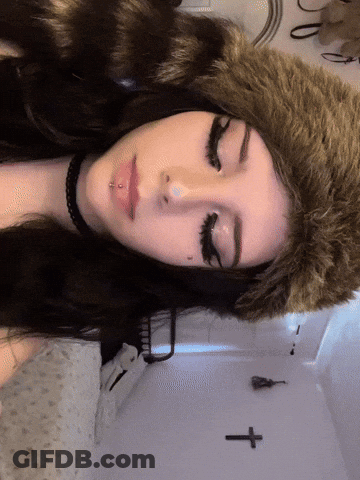A Woman Wearing A Fur Hat And A Choker Has A Cross On The Wall Behind Her GIF
