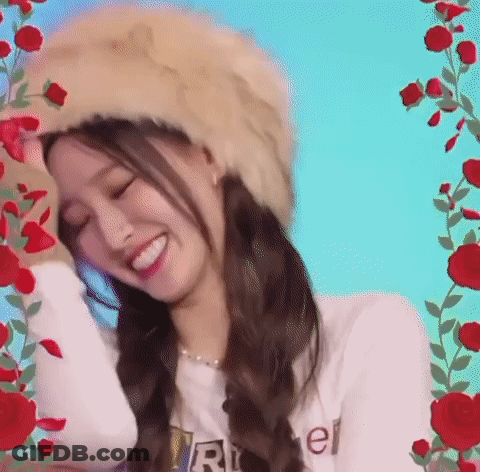 A Woman Wearing A Fur Hat And A White Shirt Is Smiling In Front Of Red Roses . GIF