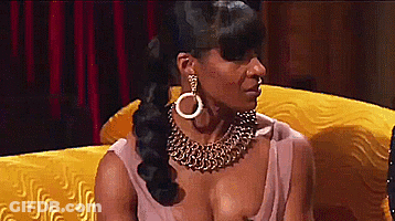 A Woman Wearing A Gold Necklace And Earrings Is Sitting On A Yellow Couch . GIF