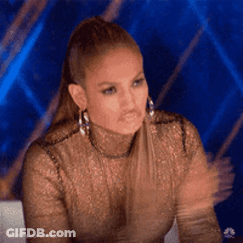 A Woman Wearing A Gold Top And Earrings Is Sitting In Front Of A Blue Background . GIF