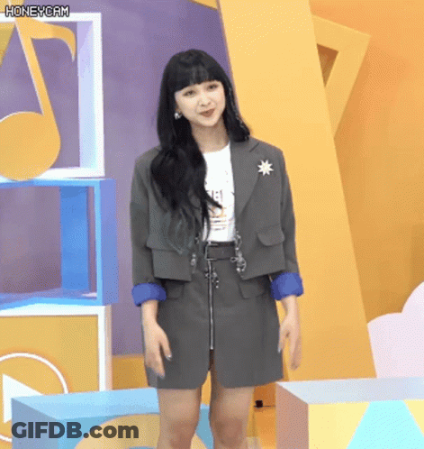 A Woman Wearing A Gray Jacket And Skirt Stands In Front Of A Yellow Wall With A Honeycam Watermark GIF