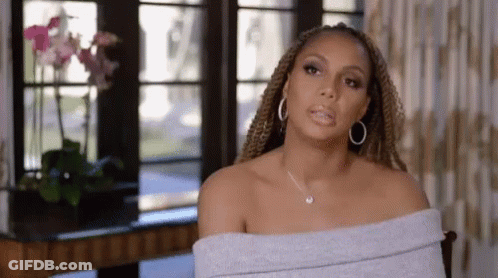 A Woman Wearing A Gray Off The Shoulder Top And Hoop Earrings Is Sitting In A Chair In Front Of A Window . GIF