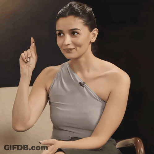 A Woman Wearing A Gray One Shoulder Top Is Pointing Upwards GIF