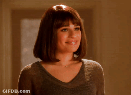 A Woman Wearing A Gray Sweater And A Necklace Smiles GIF