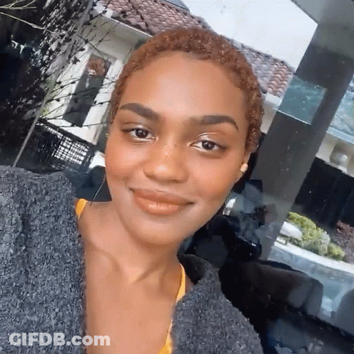 A Woman Wearing A Gray Sweater And A Yellow Top Smiles For The Camera GIF