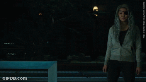 A Woman Wearing A Gray Sweater And Black Pants Stands In A Dark Room GIF