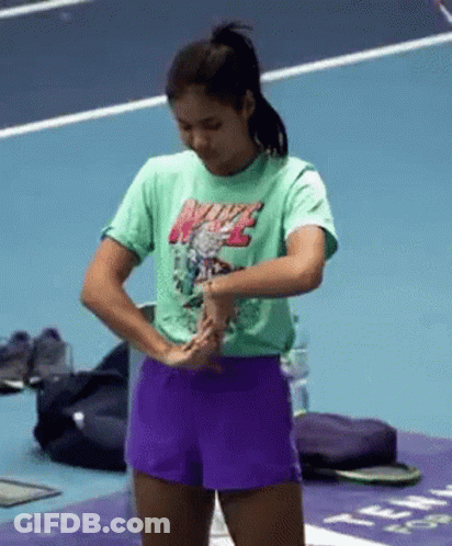 A Woman Wearing A Green Nike T-Shirt And Purple Shorts Is Standing On A Tennis Court . GIF