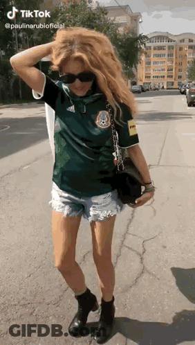 A Woman Wearing A Green Shirt And Shorts Is Standing On The Sidewalk . GIF