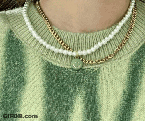 A Woman Wearing A Green Sweater And Pearl Necklaces With A Smiley Face Pendant GIF