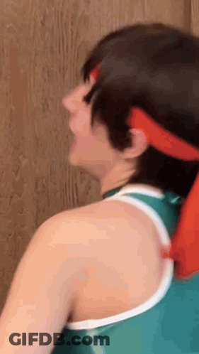 A Woman Wearing A Green Tank Top And A Red Headband Is Standing Next To A Wooden Wall . GIF