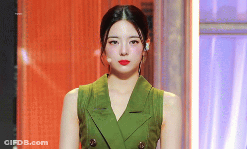 A Woman Wearing A Green Vest And Red Lipstick Is Standing In Front Of A Microphone . GIF