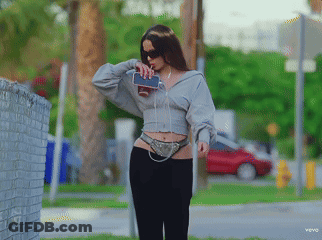 A Woman Wearing A Grey Hoodie And Black Pants Holds A Cell Phone In Her Hand GIF