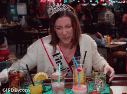 A Woman Wearing A Happy Birthday Sash Is Sitting At A Table With Drinks .. GIF