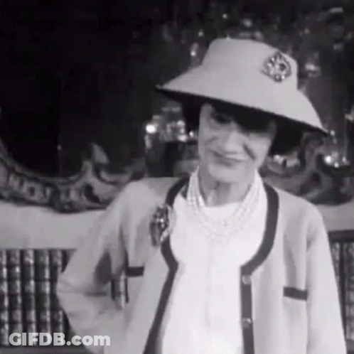 A Woman Wearing A Hat And A Cardigan Is Standing In Front Of A Bookshelf . GIF