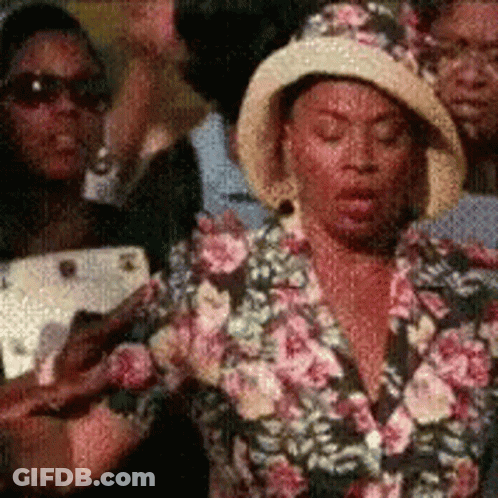 A Woman Wearing A Hat And A Floral Shirt Is Standing In A Crowd GIF