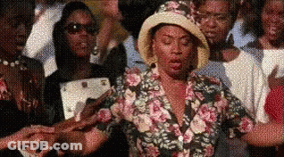 A Woman Wearing A Hat And A Floral Shirt Is Standing In Front Of A Crowd Of People . GIF