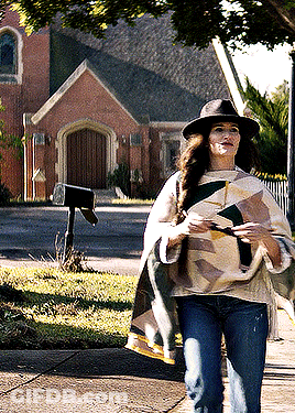A Woman Wearing A Hat And A Poncho Is Walking Down The Sidewalk GIF