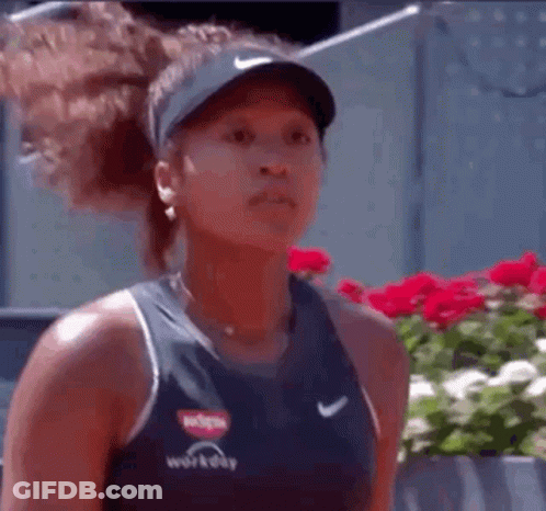 A Woman Wearing A Hat And A Tank Top Is Standing In Front Of Flowers . GIF