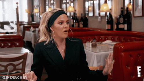 A Woman Wearing A Headband And Hoop Earrings Is Sitting At A Table In A Restaurant With The Letter E In The Corner GIF