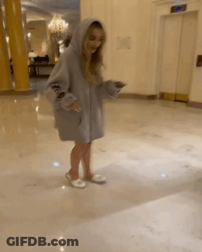 A Woman Wearing A Hoodie And Slippers Is Standing In A Hallway Next To An Elevator GIF