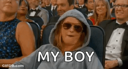 A Woman Wearing A Hoodie And Sunglasses Is Sitting In A Crowd Of People And Says `` My Boy '' . GIF