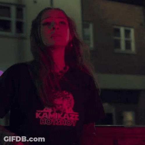 A Woman Wearing A Kamikaze Hotshot T-Shirt Stands In Front Of A Building GIF