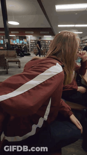 A Woman Wearing A Maroon Jacket With White Stripes On The Back Sits In A Library GIF