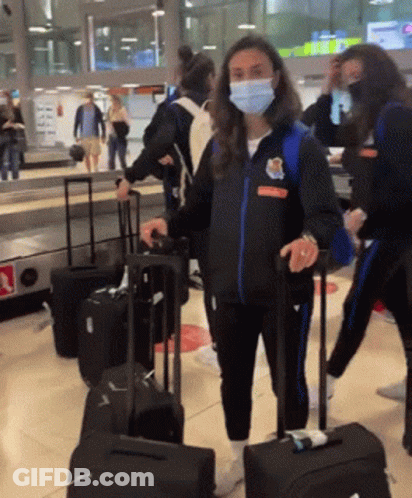 A Woman Wearing A Mask And A Jacket That Says ' Athletic ' On It Is Pulling A Suitcase GIF