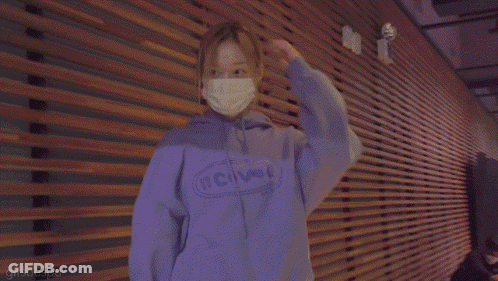 A Woman Wearing A Mask And A Purple Hoodie Is Standing In Front Of A Wooden Wall . GIF