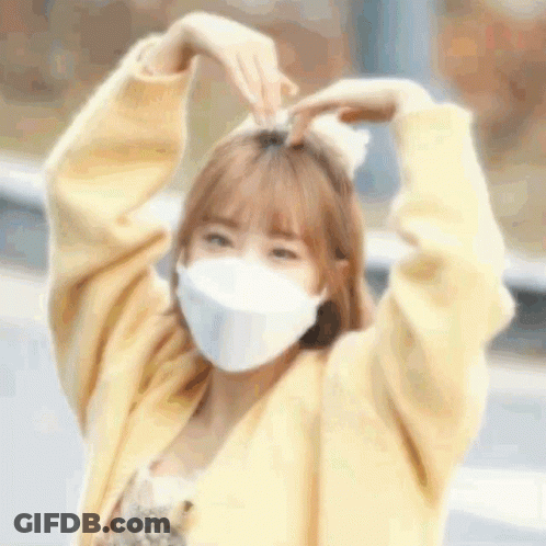 A Woman Wearing A Mask And A Yellow Sweater Is Making A Heart Shape With Her Hands . GIF