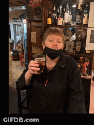 A Woman Wearing A Mask Holds A Glass Of Beer In Front Of A Sign That Says Public Telephone GIF