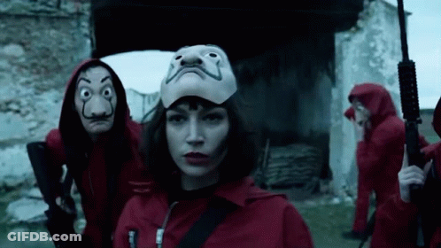 A Woman Wearing A Mask Is Standing In Front Of A Group Of People Wearing Red Jumpsuits And Holding Guns . GIF