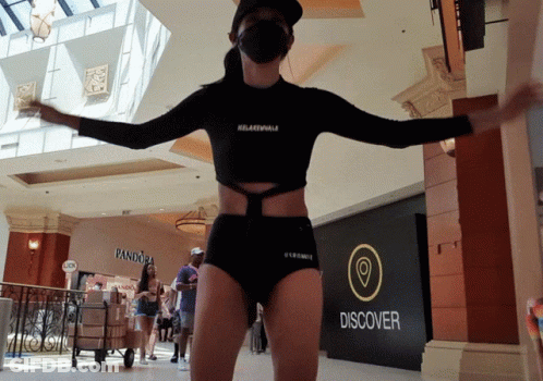 A Woman Wearing A Mask Is Standing In Front Of A Sign That Says Discover GIF