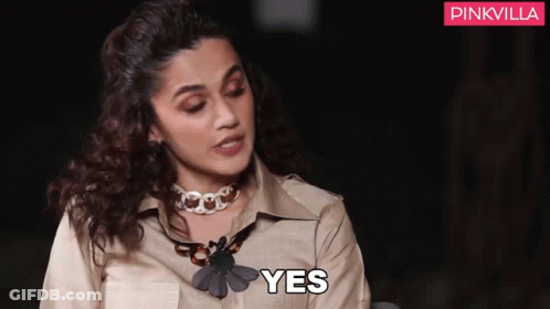 A Woman Wearing A Necklace And A Tan Shirt Is Sitting Down And Saying Yes . GIF