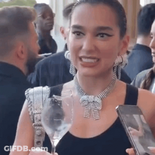 A Woman Wearing A Necklace And Earrings Is Holding A Wine Glass And A Cell Phone . GIF