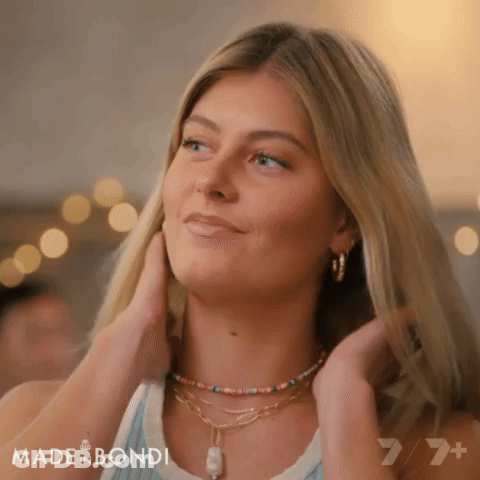 A Woman Wearing A Necklace And Earrings Is Smiling And Touching Her Hair GIF