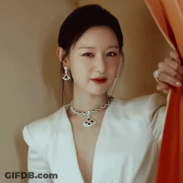 A Woman Wearing A Necklace And Earrings Is Standing In Front Of A Curtain GIF