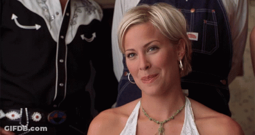 A Woman Wearing A Necklace And Hoop Earrings Is Smiling In Front Of A Man In A Cowboy Shirt GIF