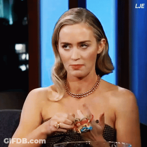A Woman Wearing A Necklace And Rings Is Sitting In Front Of A Blue Wall With The Letters Lje On It GIF