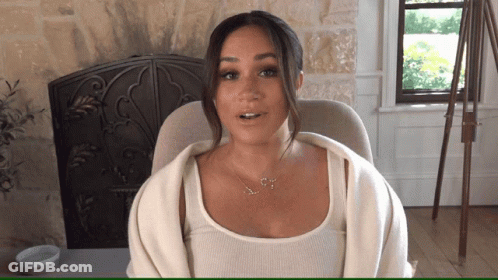 A Woman Wearing A Necklace With The Letter S On It Sits In Front Of A Fireplace GIF
