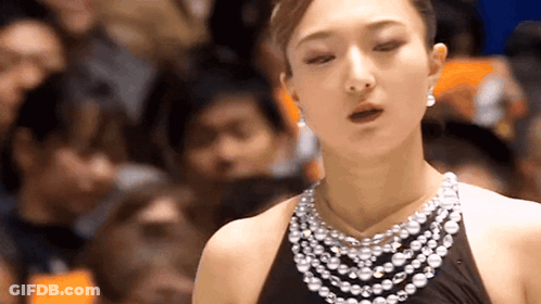 A Woman Wearing A Pearl Necklace And Earrings Is Standing In Front Of A Crowd GIF