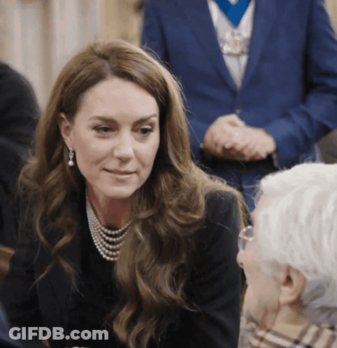 A Woman Wearing A Pearl Necklace And Earrings Talks To An Older Man GIF