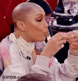 A Woman Wearing A Pearl Necklace And Hoop Earrings Is Sitting At A Table Eating A Piece Of Food . GIF