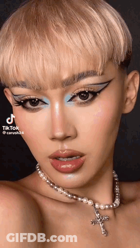 A Woman Wearing A Pearl Necklace With A Cross On It Is Looking At The Camera . GIF