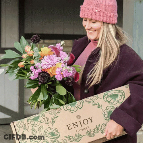 A Woman Wearing A Pink Hat Is Holding A Box Of Flowers That Says Enjoy Flowers GIF