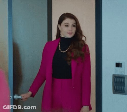 A Woman Wearing A Pink Jacket And Black Turtleneck Is Standing In A Doorway . GIF