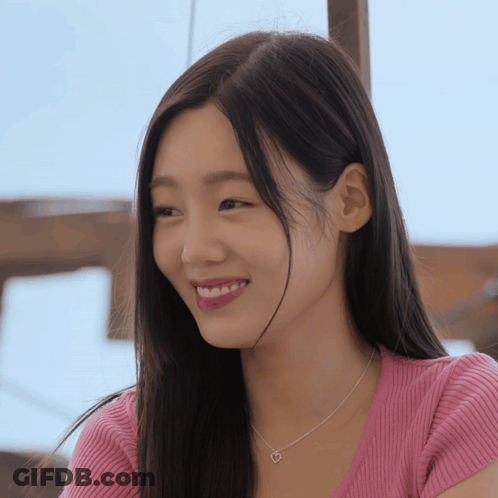 A Woman Wearing A Pink Shirt And A Heart Necklace Smiles For The Camera GIF
