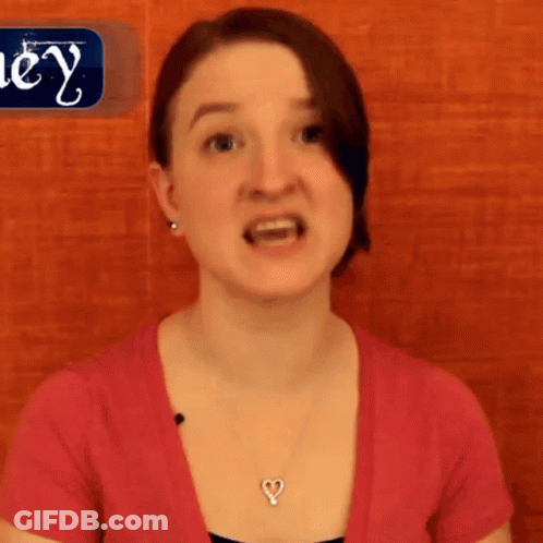 A Woman Wearing A Pink Shirt And A Heart Shaped Necklace Stands In Front Of A Sign That Says Hey GIF